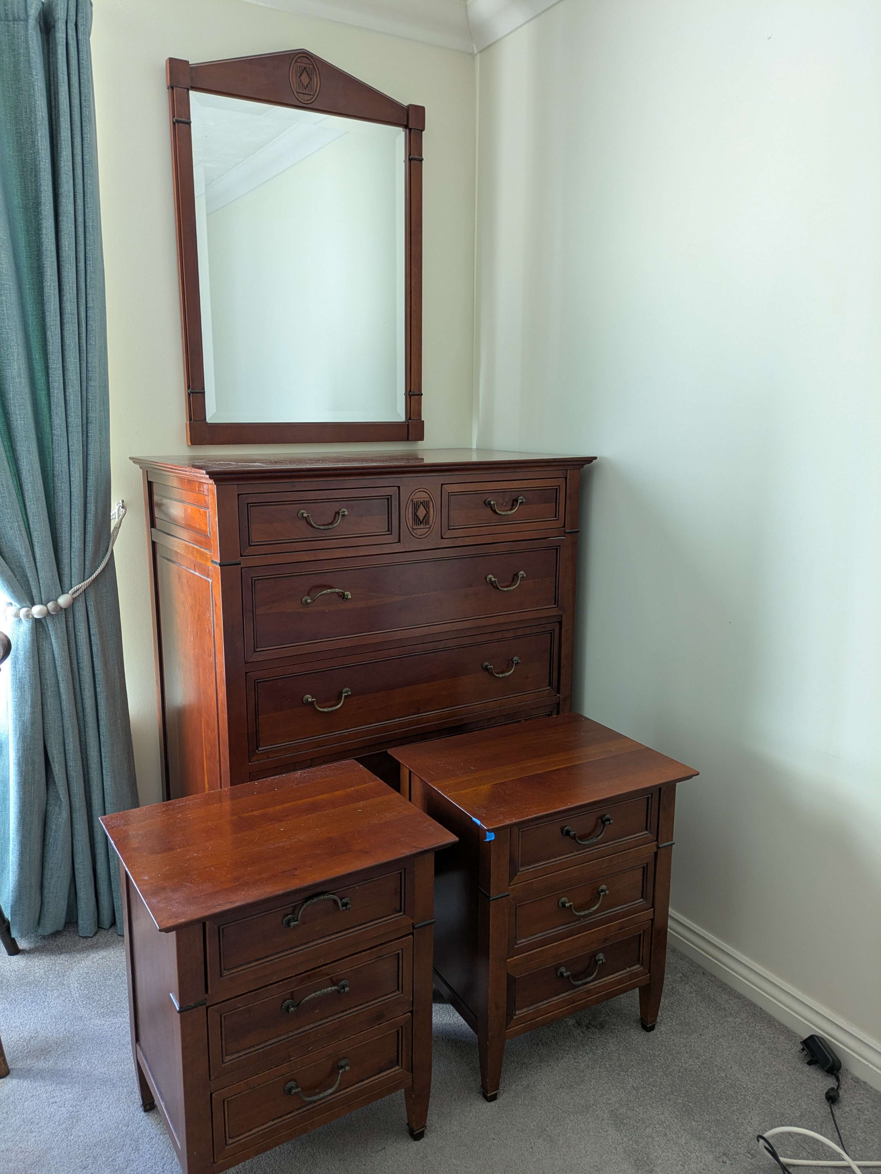 A matching mahogany bedroom set — tall chest of drawers with mirror above, and two bedside tables — left ready to move in a cream-painted bedroom